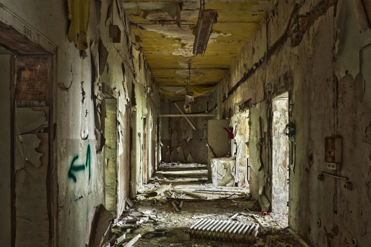 Salle d&rsquo;un b&acirc;timent abandonn&eacute;, murs nus, grandes fen&ecirc;tres et traces d&rsquo;humidit&eacute;, &eacute;voquant une ancienne zone de soins hospitaliers.