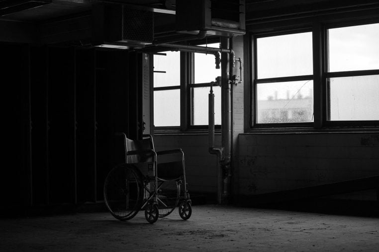 Fauteuil roulant isol&eacute; dans une pi&egrave;ce sombre d&rsquo;un ancien h&ocirc;pital abandonn&eacute;, avec de grandes fen&ecirc;tres poussi&eacute;reuses en arri&egrave;re-plan.