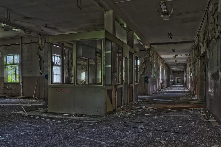 Couloir int&eacute;rieur abandonn&eacute; avec portes ouvertes et lumi&egrave;re naturelle au fond, dans un ancien b&acirc;timent hospitalier.