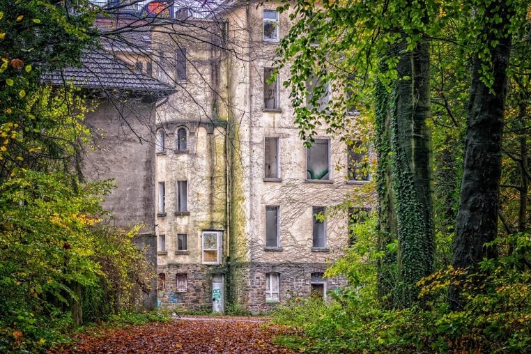 Fa&ccedil;ade d&rsquo;une clinique abandonn&eacute;e entour&eacute;e de v&eacute;g&eacute;tation, vitres cass&eacute;es et murs marqu&eacute;s par le temps.